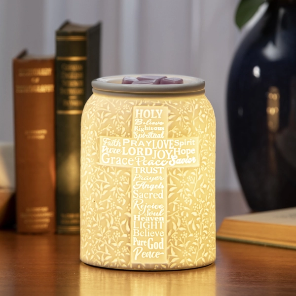 Scentsy Trust In Him Wax Warmer glowing on a wooden table, featuring pure white ceramic with an illuminated cross design made of inspirational words.