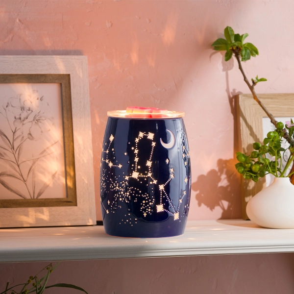 The Scentsy Constellations Electric Fragrance Melter in dark navy blue ceramic projecting illuminated star and moon patterns on a pink wall.