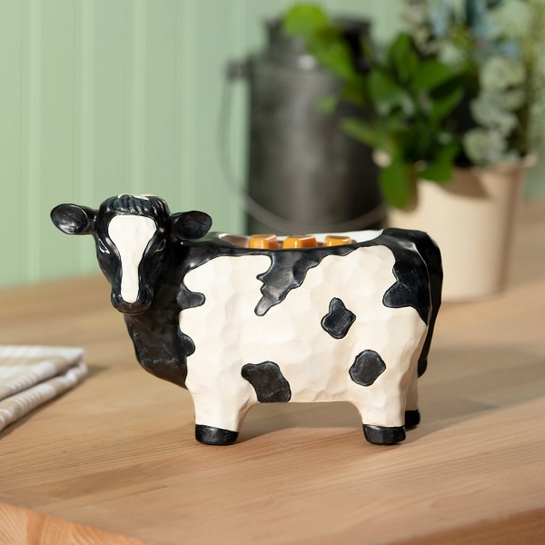The Scentsy Deja Moo Wax Warmer shaped like a black and white cow with a hand-painted, carved wood texture sitting on a table.