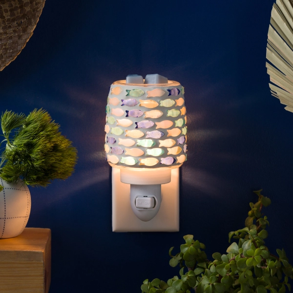 The Scentsy Fish in the sea mini Tart Warmer glowing brightly with a colorful mosaic of hand-placed glass fish on a blue wall.