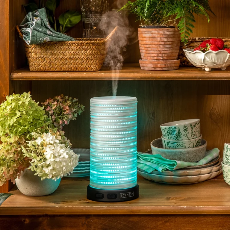 The Scentsy Gleam Diffuser glowing with cyan LED light through a white laser-cut porcelain shade on a wooden shelf.