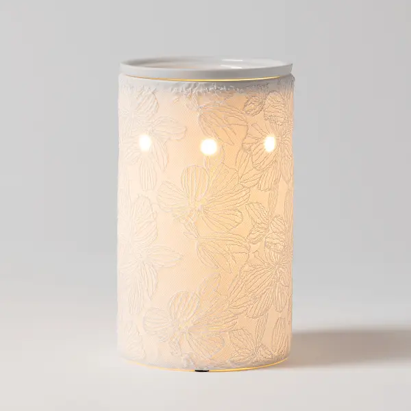 The Scentsy Lace & Light Electric Fragrance Melter features a delicate, raised floral lace pattern glowing warmly from within on a white ceramic cylinder.