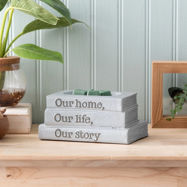 Scentsy Our Story Wax Warmer shaped like a stack of three light grey books sitting on a wooden shelf next to a plant.