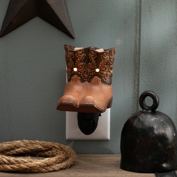 Scentsy Rodeo Mini Wax Warmer shaped like brown leather cowboy boots plugged into a wall, glowing softly from decorative cutouts.