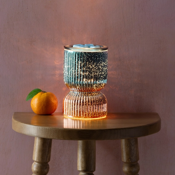 Scentsy Shine Bright Wax Warmer glowing on a table, featuring two-toned mercury glass in blue and copper with speckled light effects.