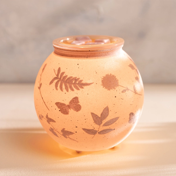 Scentsy Veiled Meadow Wax Warmer glowing on a tabletop, featuring a frosted amber glass globe with silhouettes of ferns and butterflies.